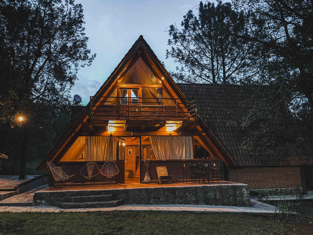 An A-frame house is illuminated at dusk, showcasing its architectural lines against a natural backdrop. The spacious porch features seating and hanging chairs, surrounded by trees, offering a cozy outdoor space. Soft light emanates from both levels, enhancing the welcoming atmosphere of the home.