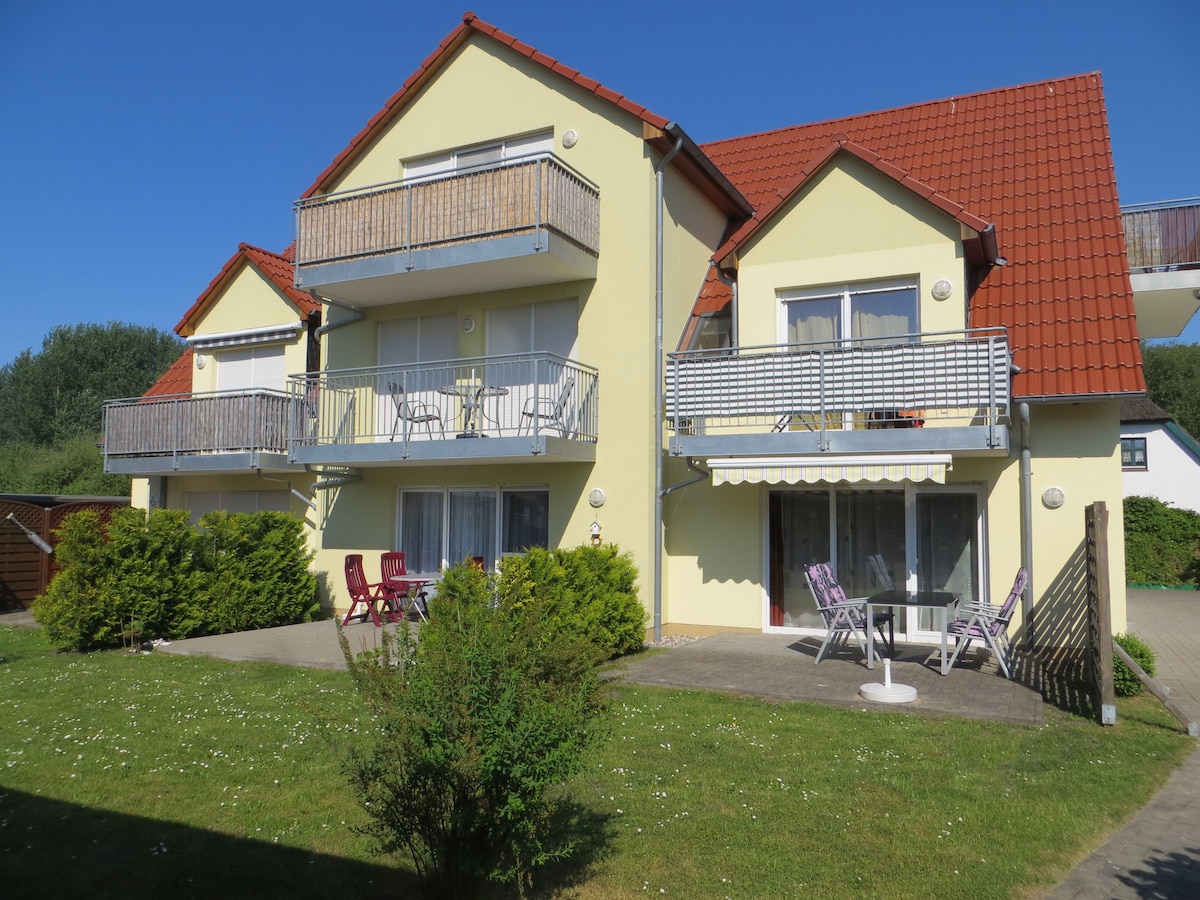 A yellow multi-family building features balconies on the upper floors, each with seating. The ground level shows a patio area with outdoor furniture. Lush greenery, including shrubbery, borders the landscaped yard, creating a pleasant environment. A clear blue sky is visible above.