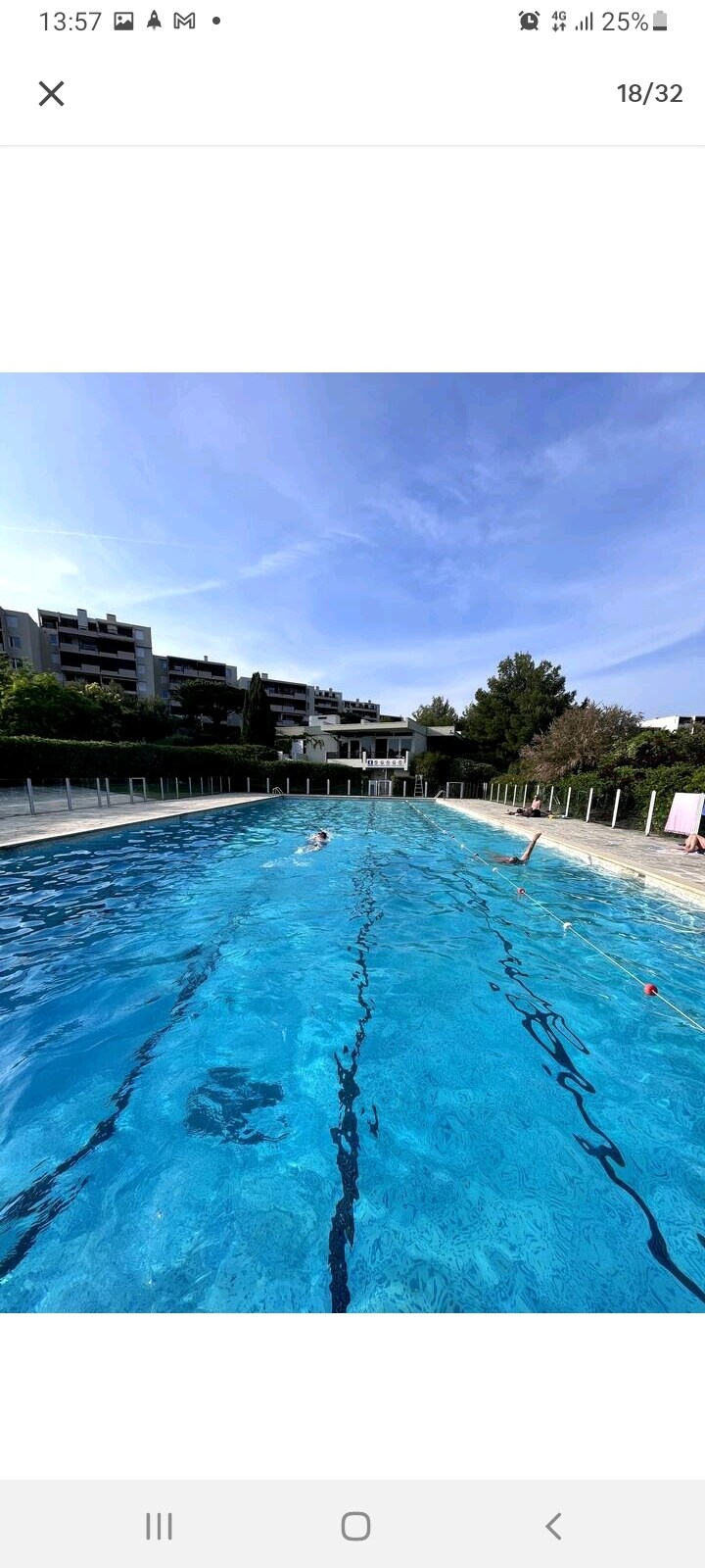 A spacious outdoor swimming pool is highlighted, featuring clear blue water under a bright sky. Surrounding the pool, green landscaping and modern residential buildings can be seen in the background, contributing to a tranquil setting. Lane markings are visible on the pool surface.