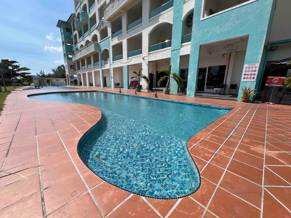 An inviting swimming pool is set in a curvy shape, surrounded by a tiled deck. The pool's clear blue water reflects the sky above. Palm trees and lounge chairs are positioned nearby, enhancing the space for relaxation and leisure.
