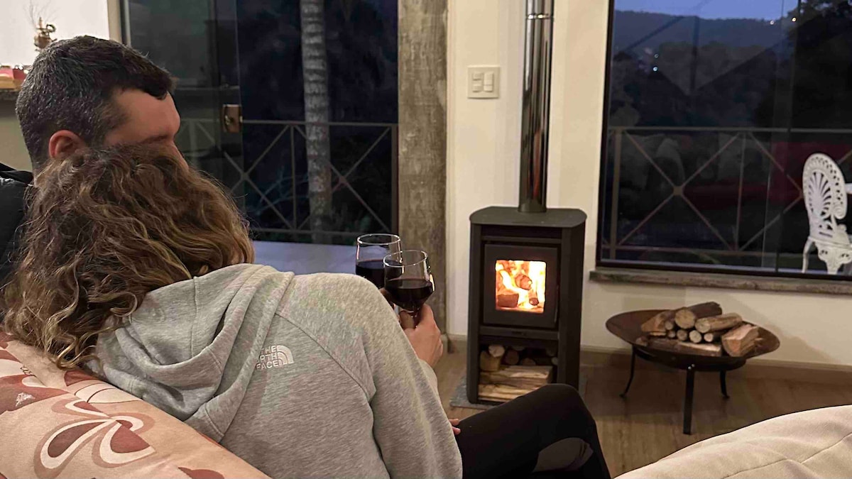 A cozy corner of the living room is displayed, featuring a small wood-burning stove emitting warmth. Soft seating is positioned nearby, and large windows provide a view of the outside landscape. A touch of elegance is added by a decorative side table.