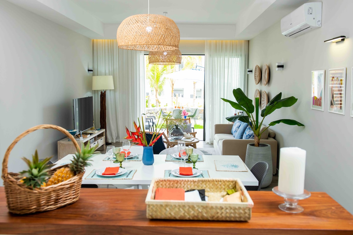 An open-plan living area is highlighted, featuring a modern dining table set for four. Tropical plants are placed strategically, and a cozy seating arrangement is visible. Natural light flows through large sliding doors, which lead to the outdoor space, enhancing the room's bright and airy atmosphere.