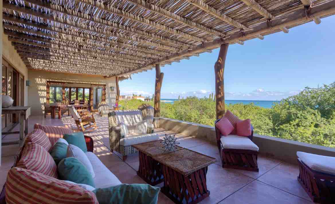 An expansive outdoor terrace is shaded by a thatched roof, featuring comfortable seating areas with colorful cushions. A coffee table made of natural materials is placed at the center, while a view of the ocean and greenery is visible in the background.