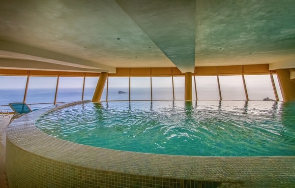 An indoor pool with a circular design is surrounded by large windows offering expansive views of the tranquil sea. The soft blue water reflects natural light, creating a serene environment. The pool area is complemented by neatly tiled edges, adding a touch of elegance.