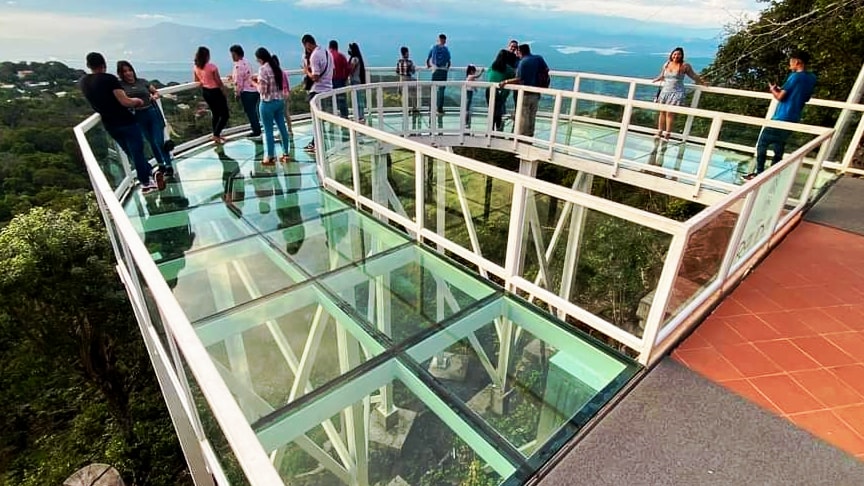 A glass observation deck is elevated among trees, offering views of surrounding landscapes. Guests are seen engaging on the deck, which features a transparent floor and a modern railing design. The area is spacious, allowing for multiple visitors to gather and enjoy the scenic surroundings.