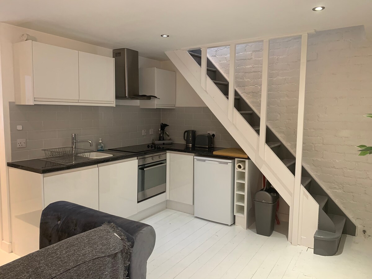 A modern kitchen area features sleek white cabinetry and stainless steel appliances, including an oven and refrigerator. Natural light brightens the space, complemented by a minimalist style. A staircase leads to the upper level, while a small plant adds a touch of greenery.