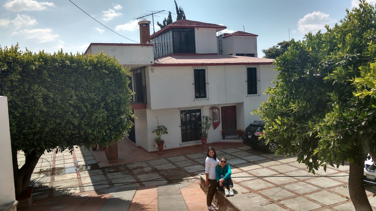 A two-story house is depicted, featuring a red-tiled roof and multiple windows. Flanking the entrance are neatly trimmed trees. A paved pathway leads toward the house, where two people are seen walking, enhancing the open and welcoming outdoor setting.