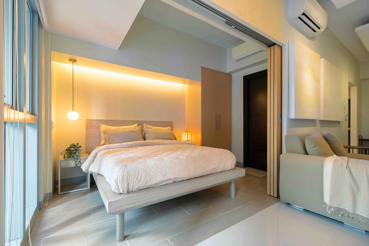 A bright and modern bedroom features a spacious bed with light linens set against a softly lit wall. A cozy seating area is positioned nearby, and large windows allow natural light to fill the space. Minimalist decor adds a sense of tranquility.