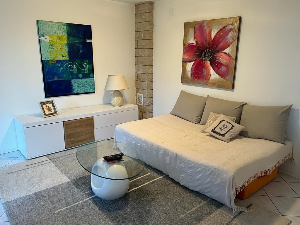 A modern living area features a white and natural wood storage unit against a wall. A round glass table sits atop a light gray rug, complementing a white couch adorned with neutral-colored cushions. Bright artwork, including a floral piece, is displayed on the walls.