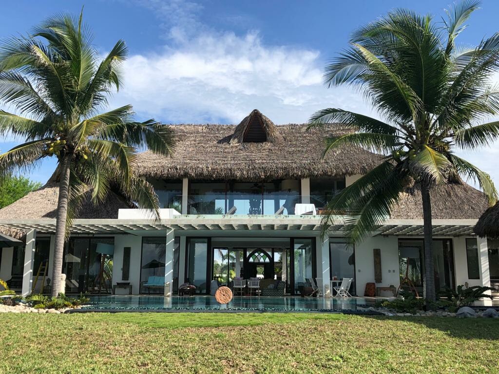 The exterior of a spacious house is framed by palm trees, presenting a thatched roof and large glass windows. A crystal-clear pool is visible in front, reflecting the sky. Lush greenery surrounds the property, contributing to a serene atmosphere.