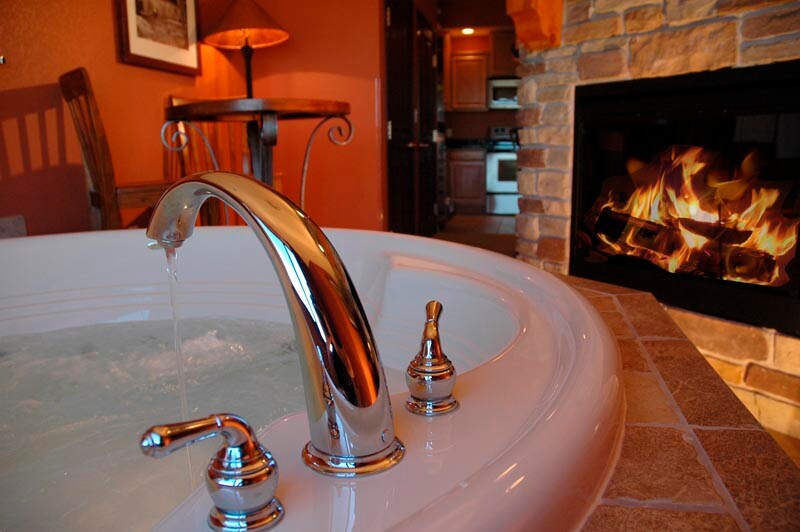 A spacious jacuzzi featuring a sleek chrome faucet is shown, with water gently flowing into the tub. A cozy indoor fireplace, framed by warm stone, is visible in the background, creating a relaxing ambiance in the studio condominium.