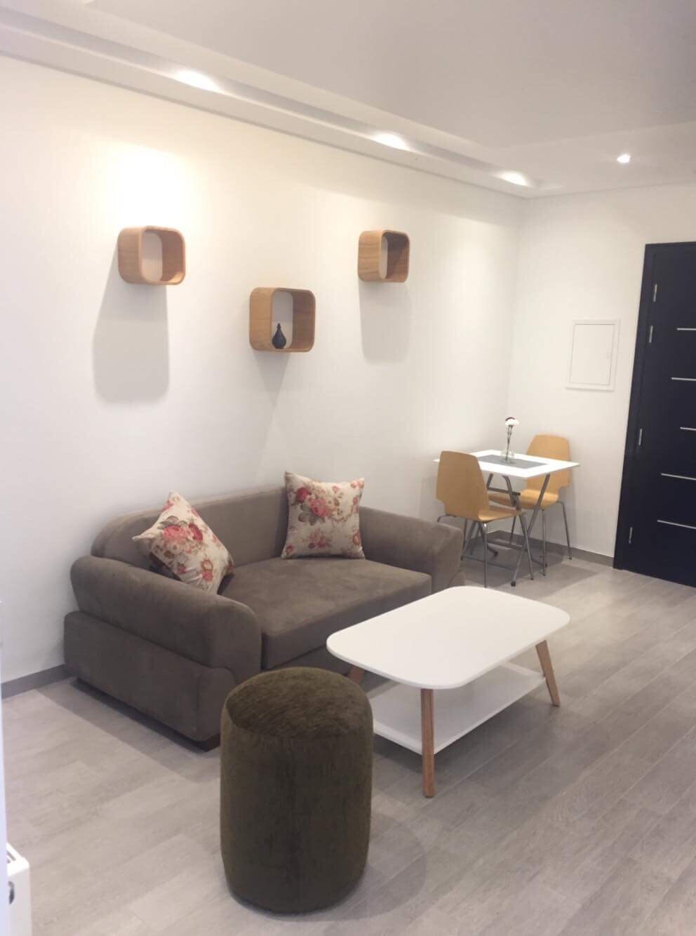 A modern living space features a plush sofa adorned with floral-patterned cushions. A minimalist coffee table sits in front, while two wooden shelves are mounted on the wall. A dining area with a small table and two chairs is visible in the background.