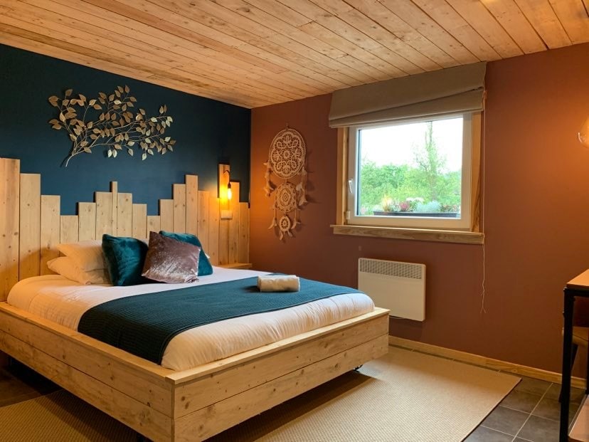 A cozy bedroom features a spacious bed with a dark blue blanket and soft pillows. Natural wood accents create warmth, while a large window provides natural light and views of greenery. Artwork on the wall adds a decorative touch, complementing the serene atmosphere.