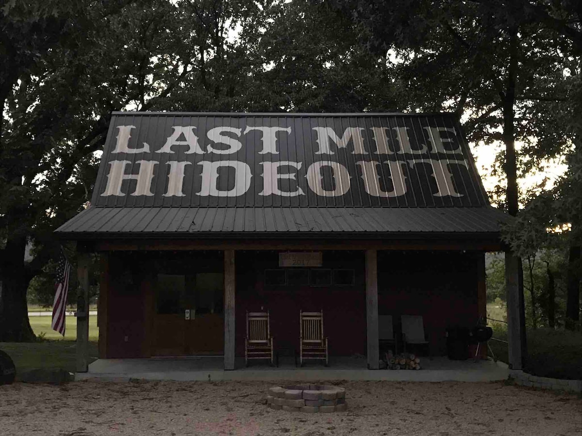 The exterior of The Last Mile Hideout is displayed with a large, prominent sign on the metal roof. The front porch features two rocking chairs, welcoming guests to enjoy the surrounding wooded area. Soft evening light enhances the tranquil setting.