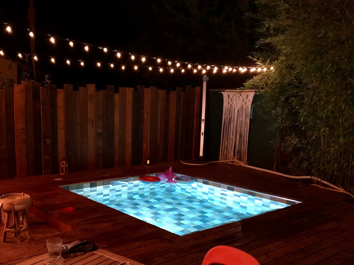 A private pool is set on a wooden deck, with colorful tiles reflecting soft lights. Surrounding greenery adds a sense of seclusion. String lights overhead create a warm ambiance, complementing the inviting atmosphere of the outdoor space.