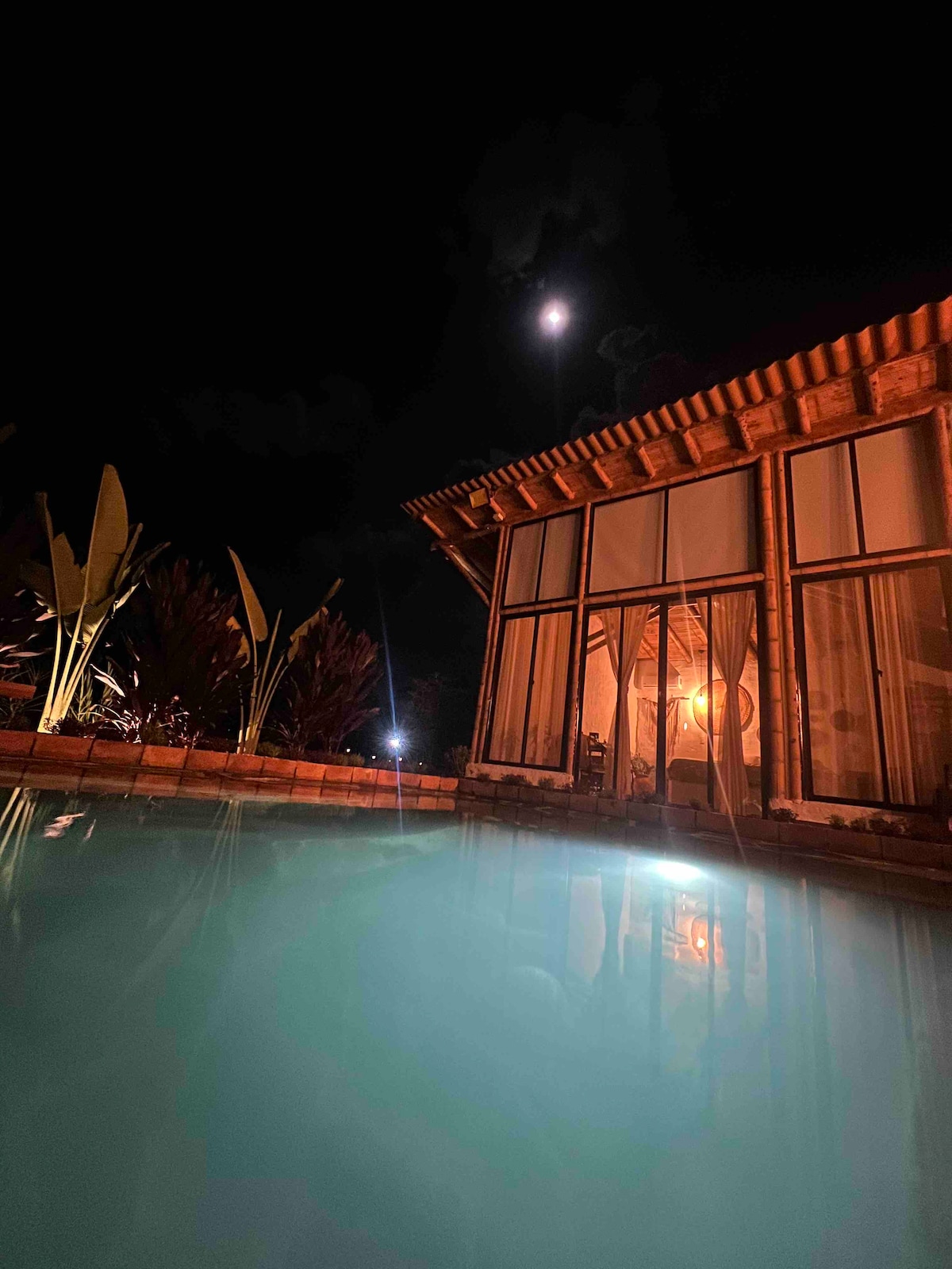 A serene nighttime view of the property shows a softly illuminated house with large glass windows reflecting the glow of the moon. The still waters of the nearby pool mirror the tranquil ambiance, while lush greenery surrounds the area, enhancing the peaceful setting.