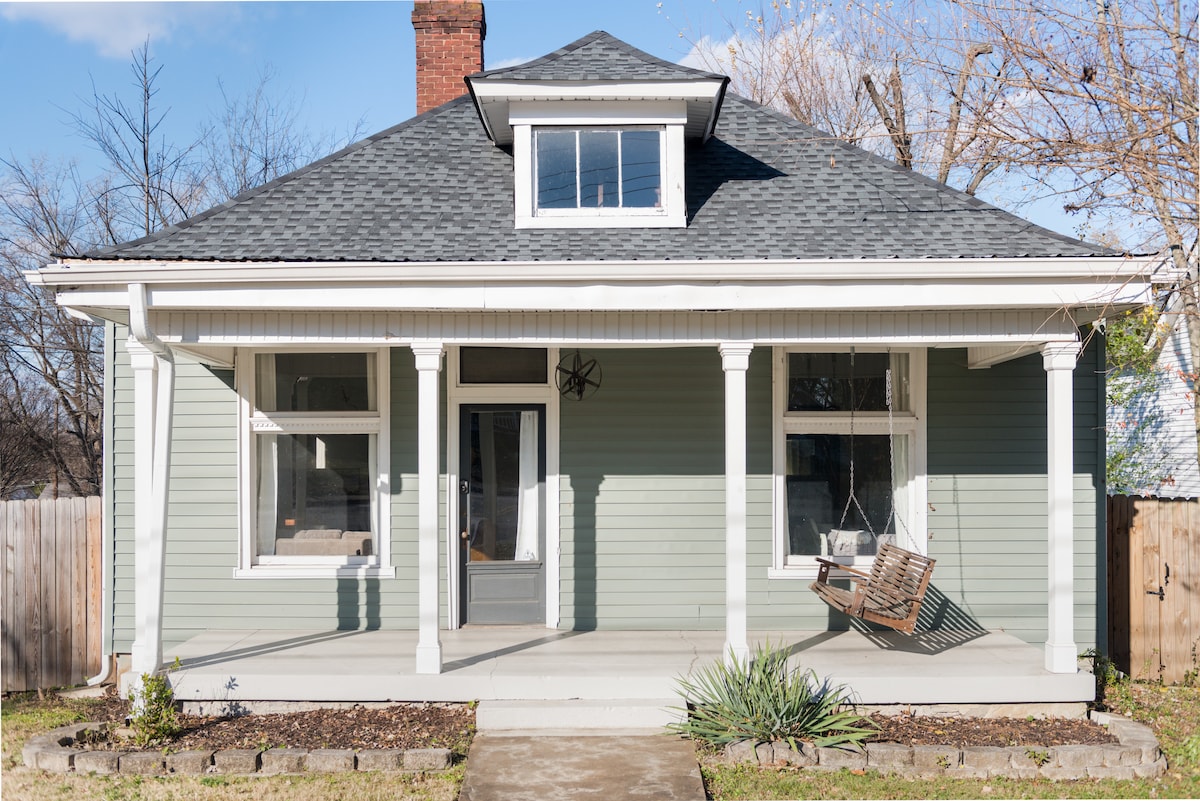 The exterior of a remodeled bungalow is displayed, featuring a welcoming front porch with a swing and two large windows on either side of the entrance. The soft green siding complements the white trim, while a small garden area with rocks and plants adorns the front yard.