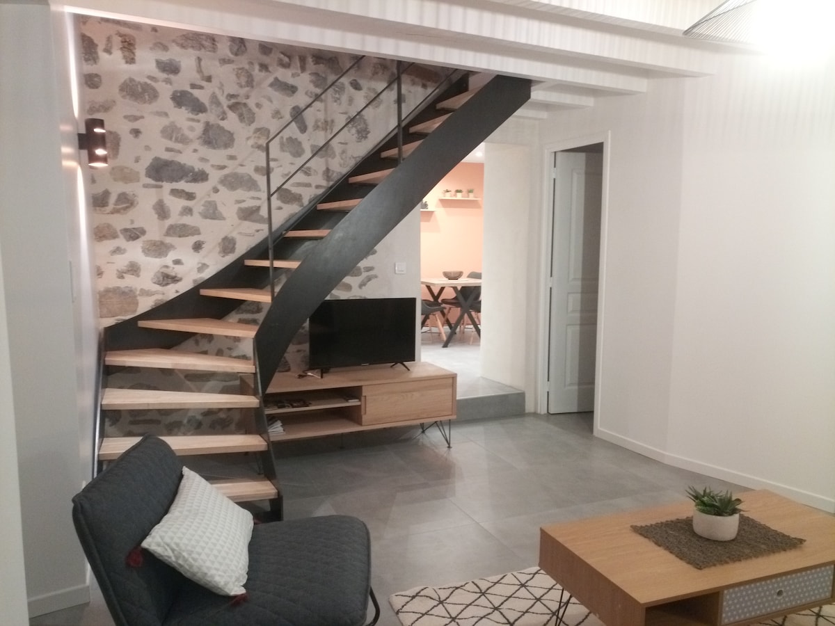 A modern living area features a stylish spiral staircase made of wood and metal, juxtaposed against an exposed stone wall. A sleek TV is mounted on a wooden console. A cozy armchair and a small table with a decorative centerpiece complete the space, illuminated by soft lighting.