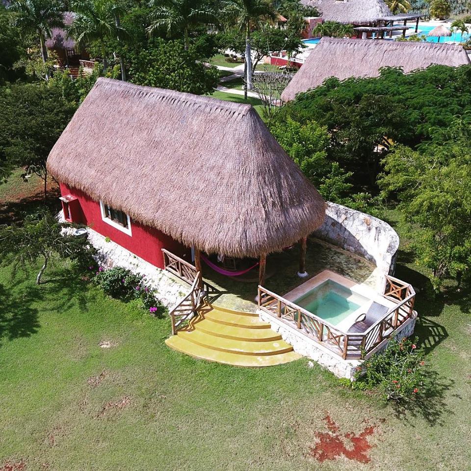 A spacious cabin with a thatched roof is set in a lush green landscape. A private hot tub is located on a stone terrace, surrounded by tropical plants. The cabin features a vibrant red exterior, providing a warm contrast to the natural surroundings.