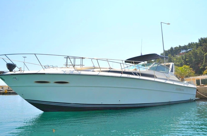 A sleek white motorboat is anchored in calm waters, reflecting the clear blue sky. The boat's design features a spacious deck with a canopy for shade, and its hull showcases subtle lines that enhance its elegant appearance.