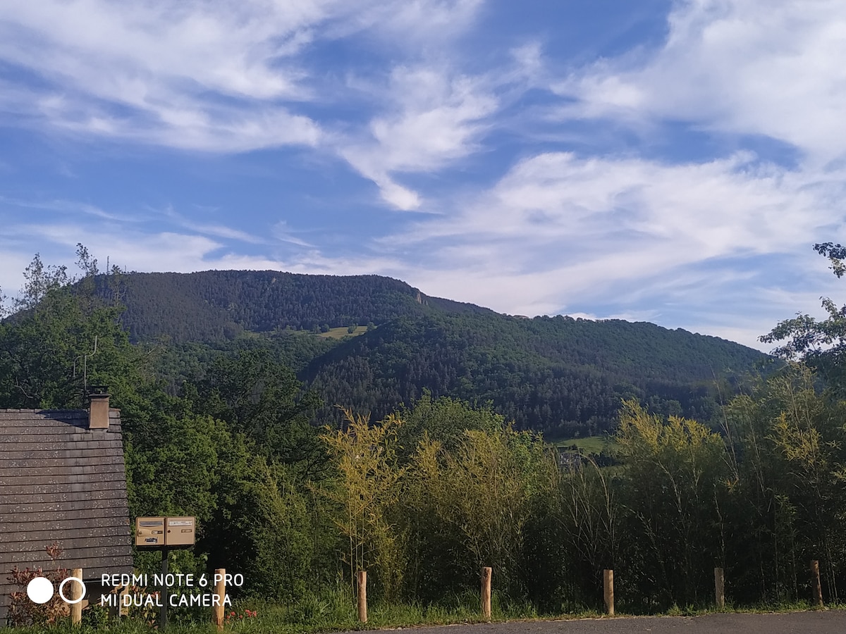 A scenic view of a mountainous landscape, showcasing lush green hills under a partly cloudy sky. The foreground features natural greenery and wooden posts, suggesting a tranquil outdoor environment. The grandeur of the mountains adds to the serene setting.