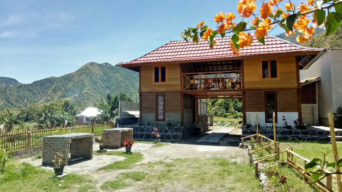 A two-story wooden house is depicted against a backdrop of rolling green hills and a clear sky. The welcoming exterior features a red-tiled roof and an open balcony, while a pathway leads to the entrance. Colorful flowers add a touch of nature to the surroundings.