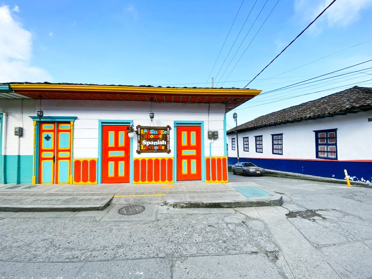 The exterior of a colorful house features vibrant doors and window frames, representing the local culture. A decorative sign is visible above the entrance. The building is adjacent to a well-maintained street, with power lines in the background and a parked car nearby.
