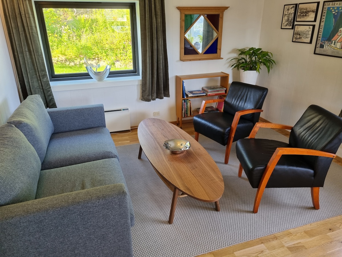 A cozy living area is furnished with a gray sofa, two black armchairs, and a wooden coffee table. Natural light enters through the large window, illuminating the space. A plant adds a touch of greenery, and shelves are visible in the background, showcasing books and decorative items.