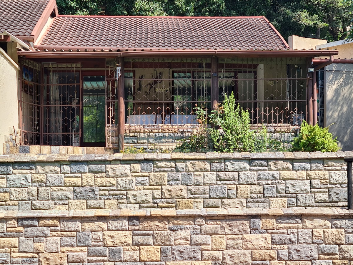The image presents a charming exterior view of a single-unit residence. A stone wall surrounds the property, with an intricate metal gate leading to a covered patio adorned with greenery. The unit is topped with a tiled roof and features large windows for natural light.