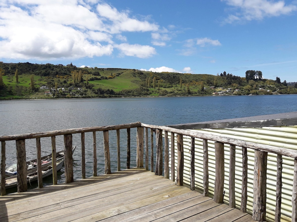A wooden terrace extends over the water, offering views of rolling green hills and a calm river. Low vegetation and a few scattered homes can be seen in the distance, under a partly cloudy blue sky.
