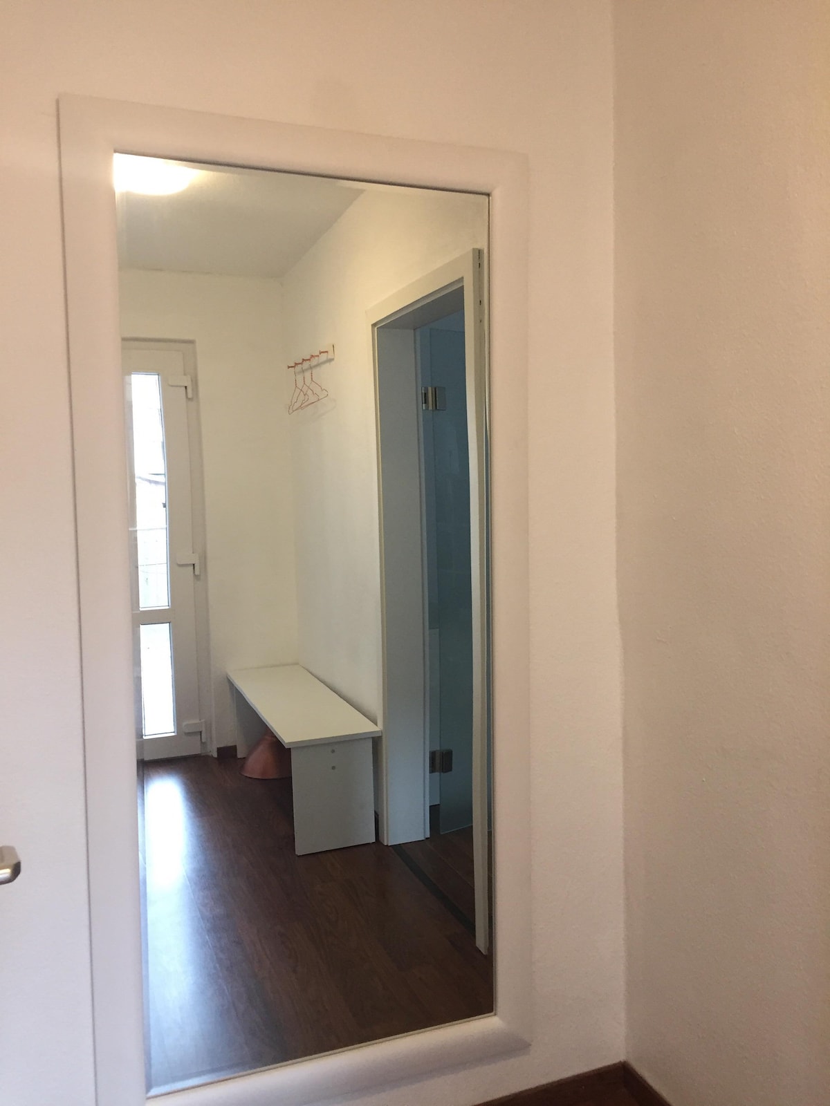 A bright entryway features light-coloured walls and a wooden floor. A large mirror is mounted on the wall, reflecting the space, while a bench provides seating. A door leads to the outside, and hanging hooks are visible nearby.