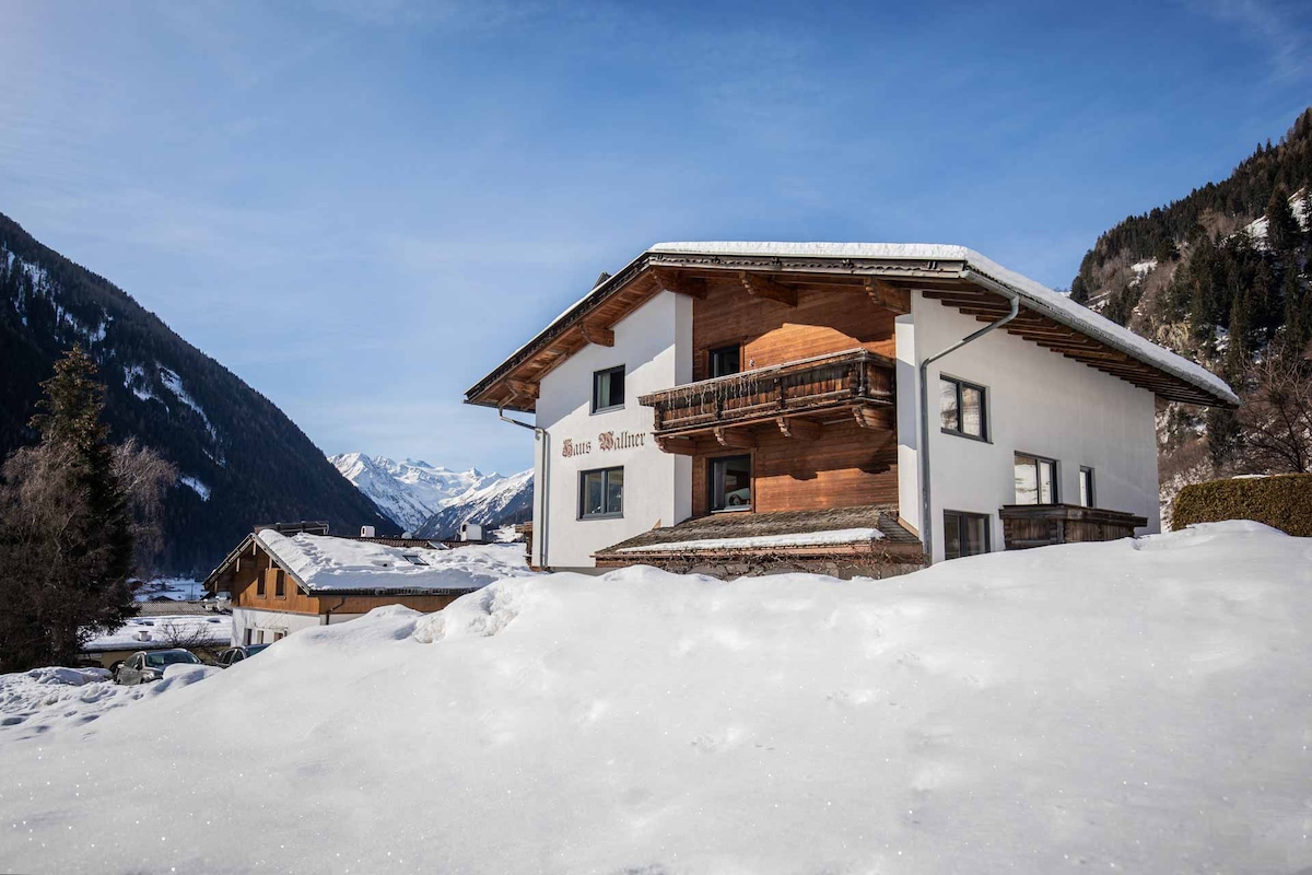 The exterior of Haus Wallner is displayed, featuring a two-story building with a combination of wood and plaster elements. Snow blankets the ground, and the surrounding mountains are visible in the distance under a clear blue sky.