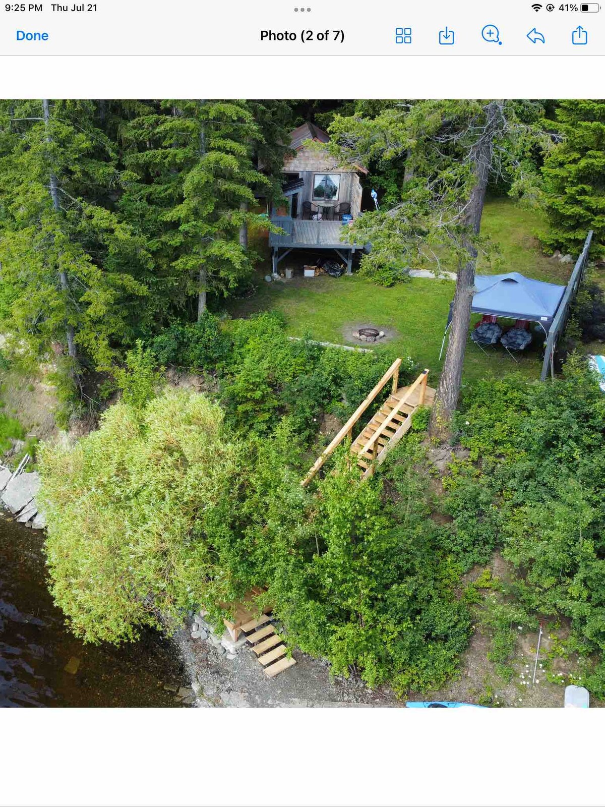 An aerial view captures a cozy cabin surrounded by lush greenery, situated near the water's edge. A wooden staircase leads down to the shoreline, highlighting a rustic fire pit area among trees. A shaded gazebo is visible nearby, completing the serene lakeside setting.