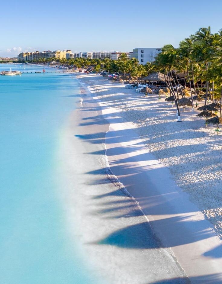 A tranquil beach scene features soft white sand meeting crystal-clear turquoise waters. Shadows from palm trees stretch along the shoreline, creating a serene ambiance. In the distance, vacationers can be seen enjoying the beach, while modern buildings frame the background against a clear blue sky.