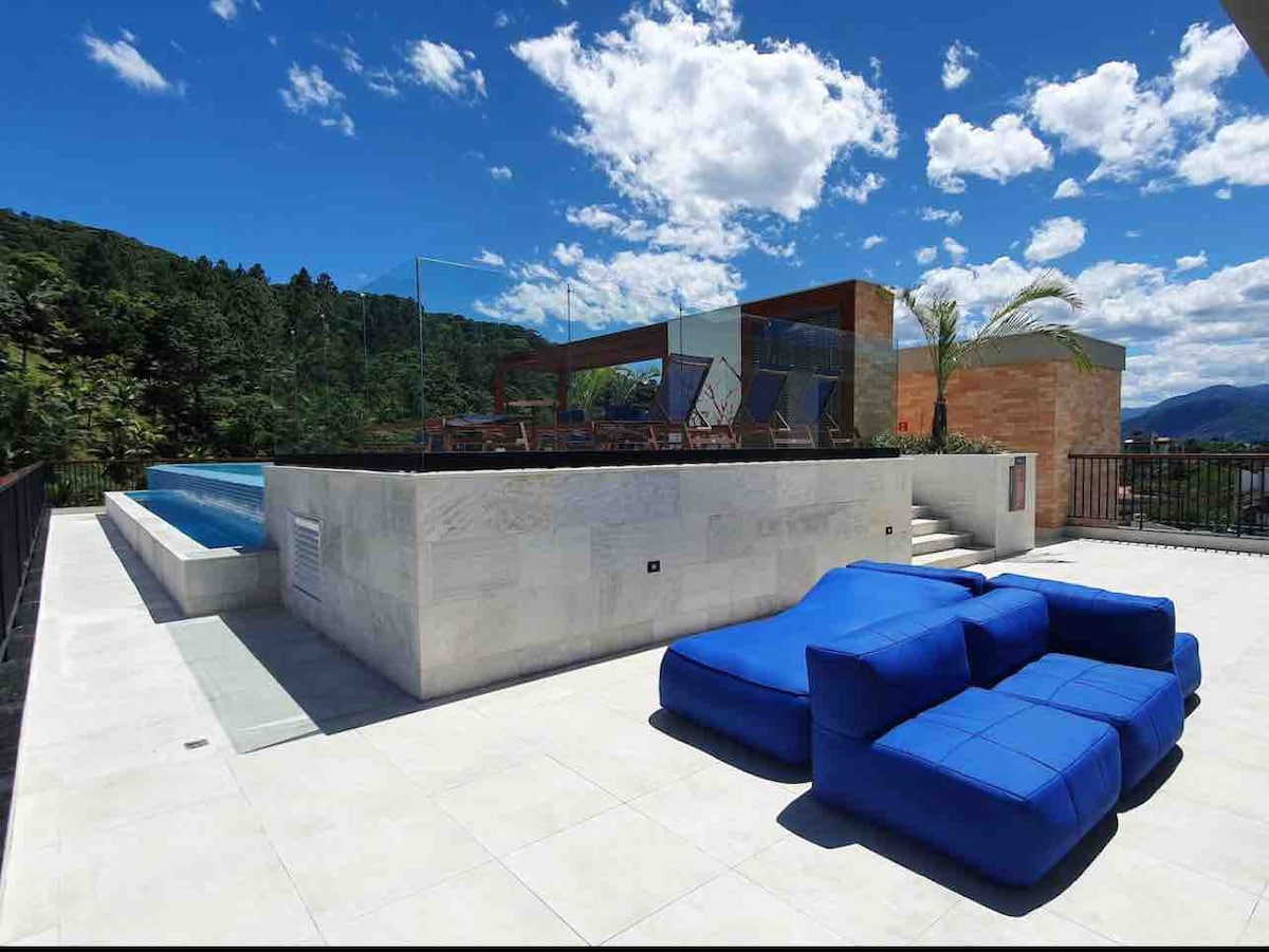 An outdoor terrace displays a bright blue sectional sofa alongside a shimmering pool. The area is surrounded by glass railings, with shaded seating and dining options visible in the background. Lush greenery frames the space under a clear blue sky with scattered clouds.