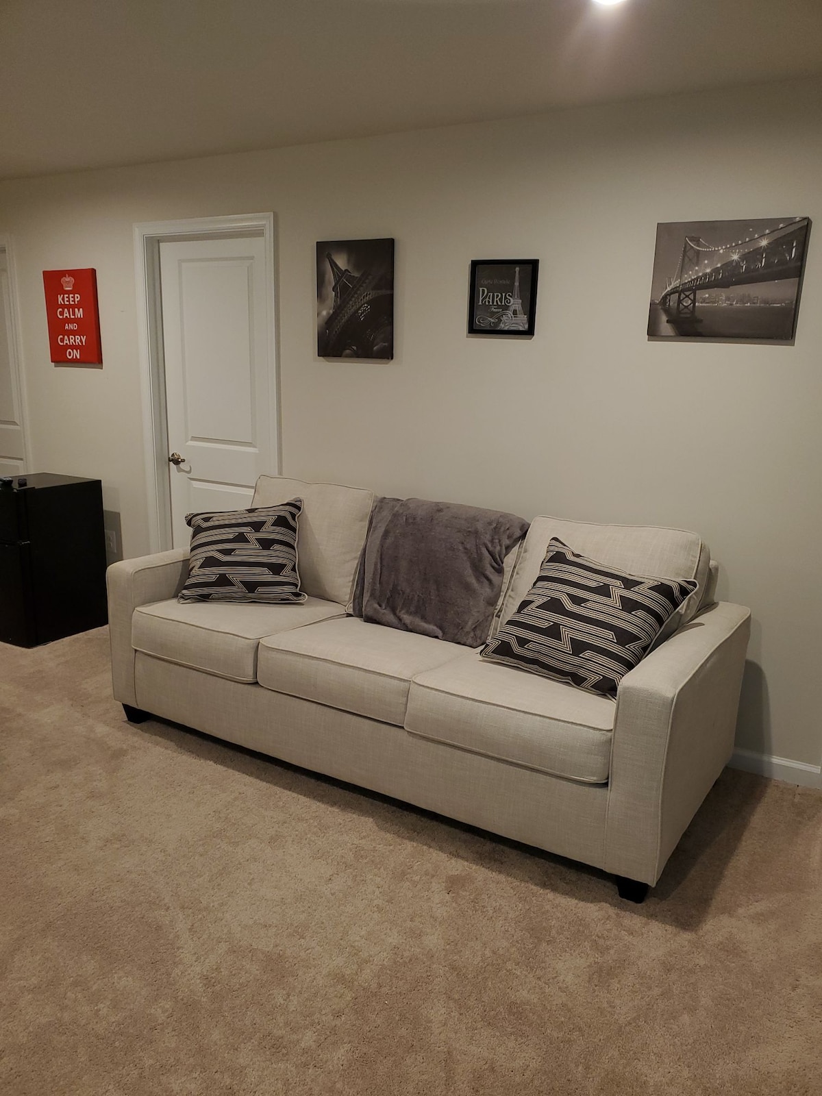 A light-colored sleeper sofa is positioned against a neutral wall, adorned with two decorative pillows. Above, three black and white framed photographs are displayed. A cozy throw is casually draped over the arm of the sofa, while a small cabinet is visible in the background.