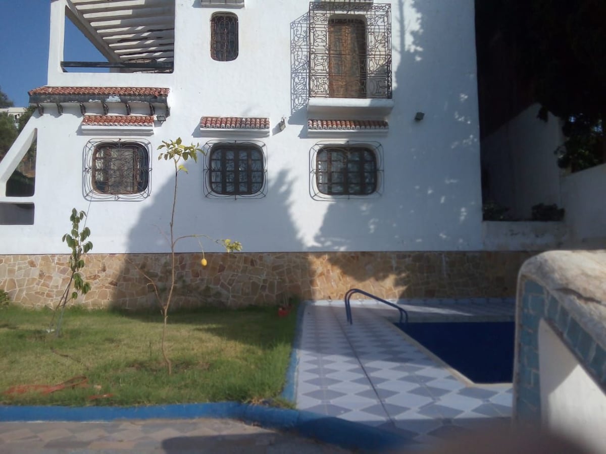 A charming exterior is depicted, showcasing a white structure with delicate wrought iron windows. A small green tree is positioned in the foreground, while a rectangular swimming pool is present, surrounded by patterned tiles. The sun casts a warm light on the scene.