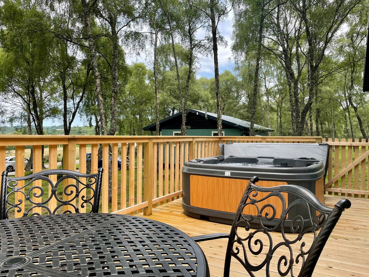 A private hot tub is situated on a wooden deck, surrounded by a wooden railing. A round table with intricate metalwork and two matching chairs is visible, providing a comfortable outdoor seating area. Lush greenery of silver birch trees frames the scene, enhancing the natural setting.