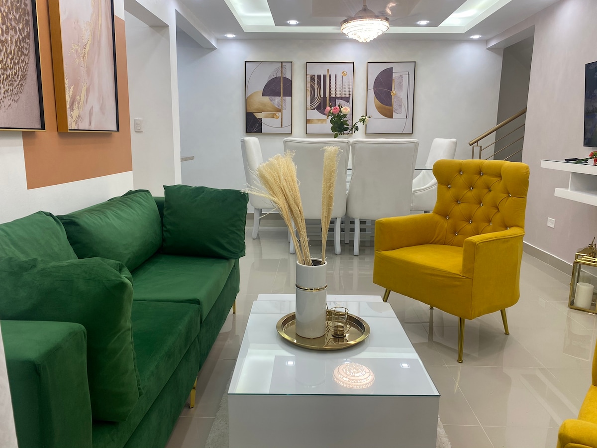 A welcoming living area features a green sofa and a yellow accent chair. A glass coffee table is placed in the center, surrounded by decorative items. Two framed artworks hang on the walls, and a dining area with white chairs is visible in the background.