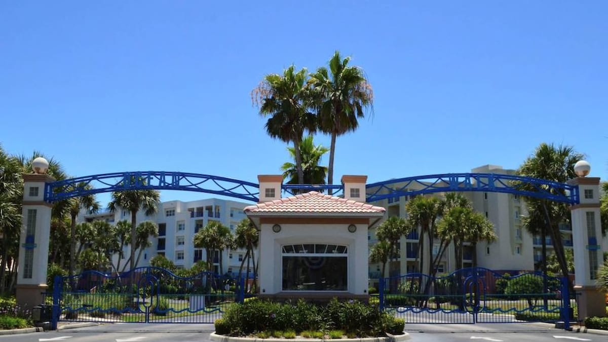 The entrance to a gated community is framed by vibrant palm trees under a clear blue sky. Blue gates create an inviting entrance, leading towards well-maintained landscaping and buildings that offer a glimpse of the serene atmosphere of the resort-style environment.