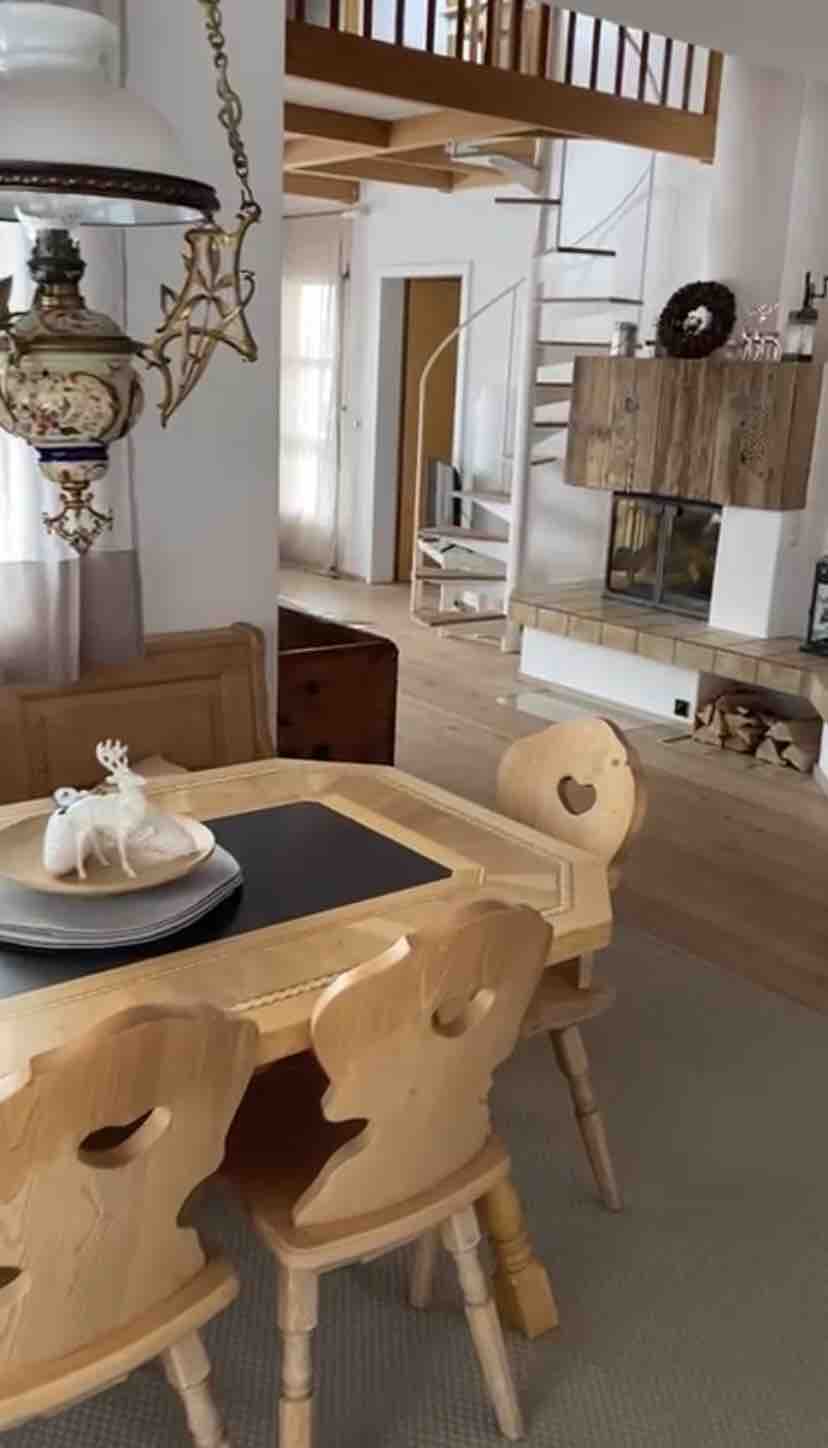 A wooden dining table is surrounded by four chairs with heart-shaped cutouts. A decorative centerpiece rests on the table. In the background, a spiral staircase leads to an upper level, and a modern fireplace is visible along the wall.