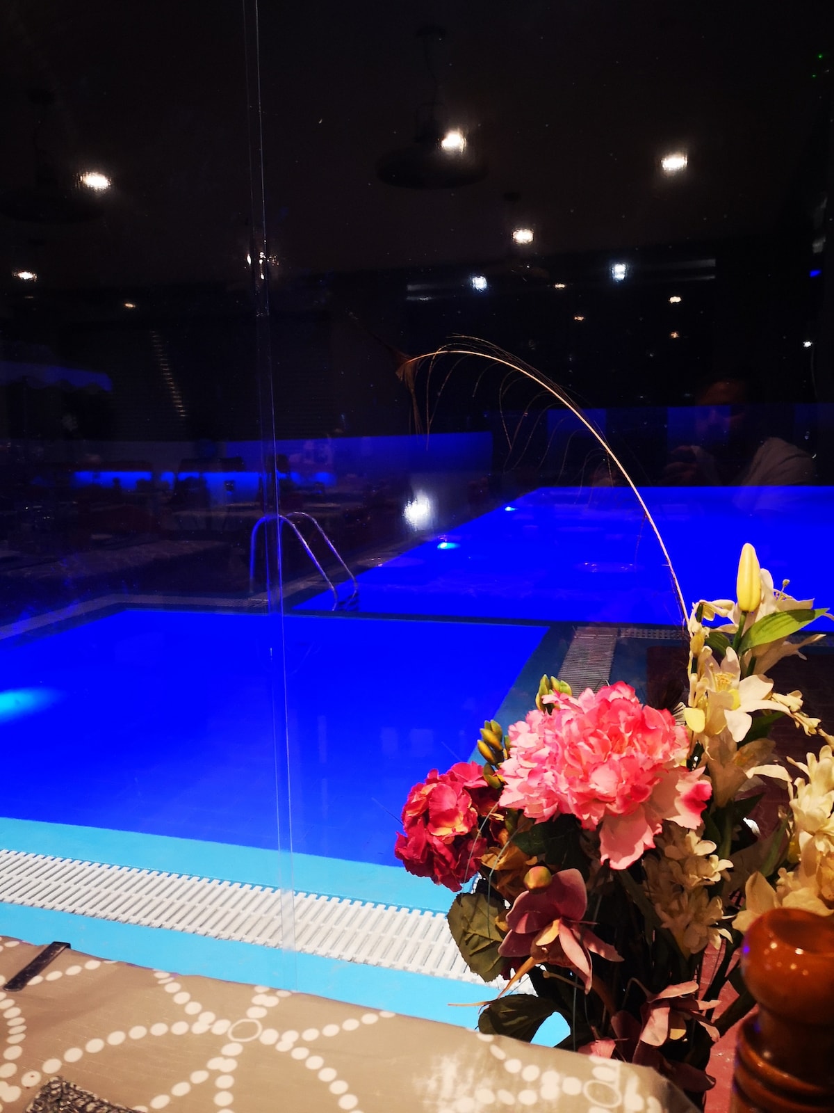 A view of a swimming pool is presented, featuring illuminated blue water that contrasts against a dark background. In the foreground, a decorative arrangement of colorful flowers adds a touch of nature, while subtle lighting enhances the inviting atmosphere.