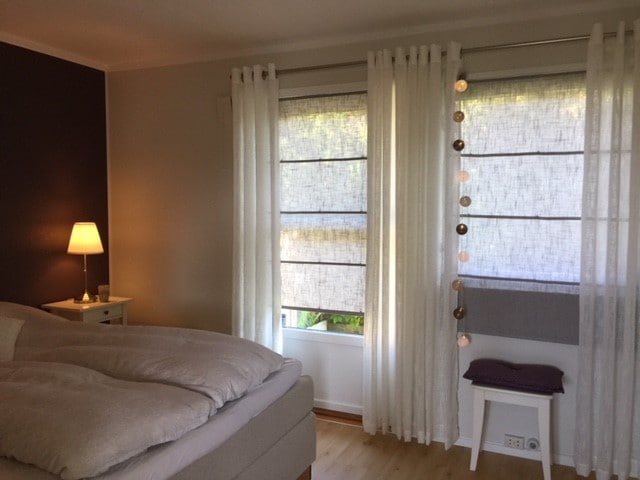 A serene bedroom features a spacious bed with plush bedding, complemented by soft white curtains framing a window. A bedside lamp provides warm lighting, while decorative string lights add a subtle touch. A small side table displays a cozy throw pillow.