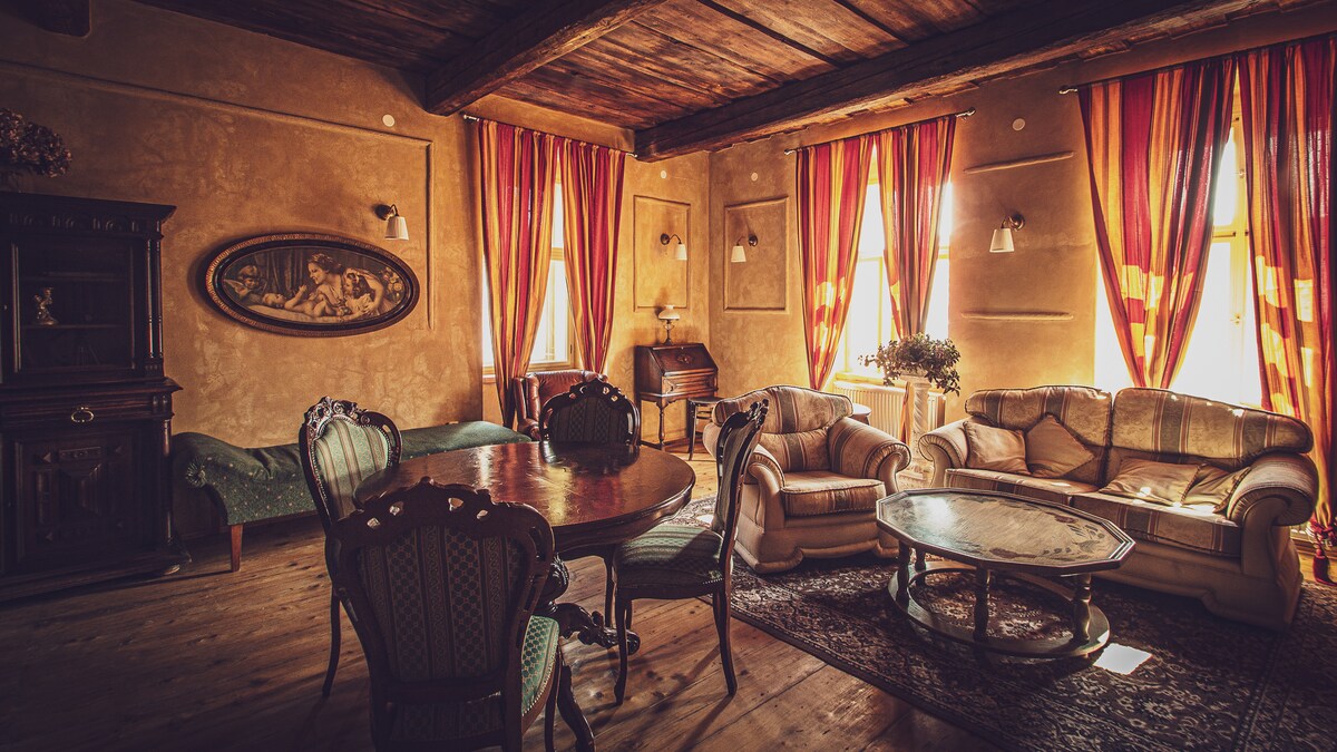 A spacious living area features rich wooden beams overhead and warm-toned walls. Elegant seating arrangements, including a plush sofa and chairs, are grouped around a central coffee table. Natural light filters through large windows, complemented by bright, flowing drapes.