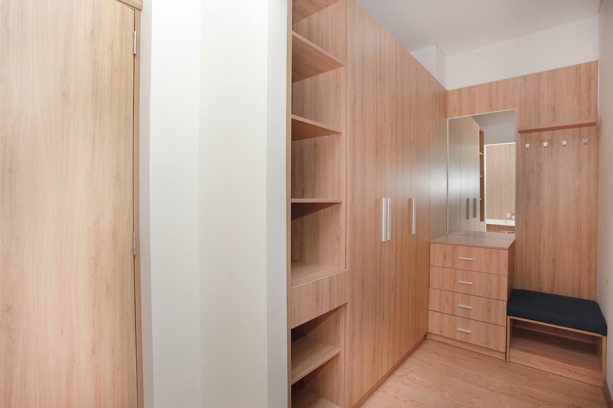 The spacious closet area features light wood paneling, with built-in shelves and drawers for storage. A full-length mirror is positioned above a low countertop, complemented by a cushioned bench. Wall hooks are available for hanging items, creating an organized and functional space.