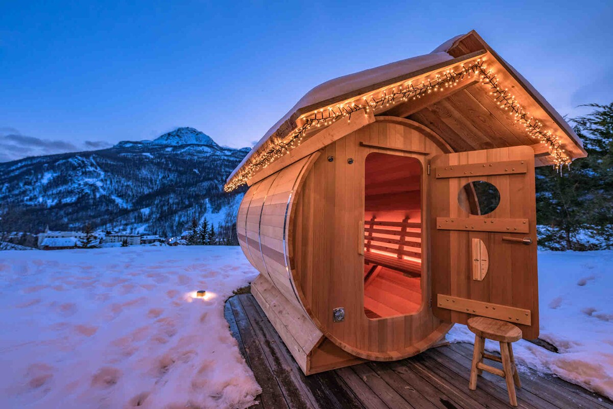 A wooden sauna sits on a clear path, illuminated by warm lights along the roof. The structure features a circular design with a large door partially open, revealing wooden benches inside. A snowy landscape extends in the background, framed by mountain peaks.