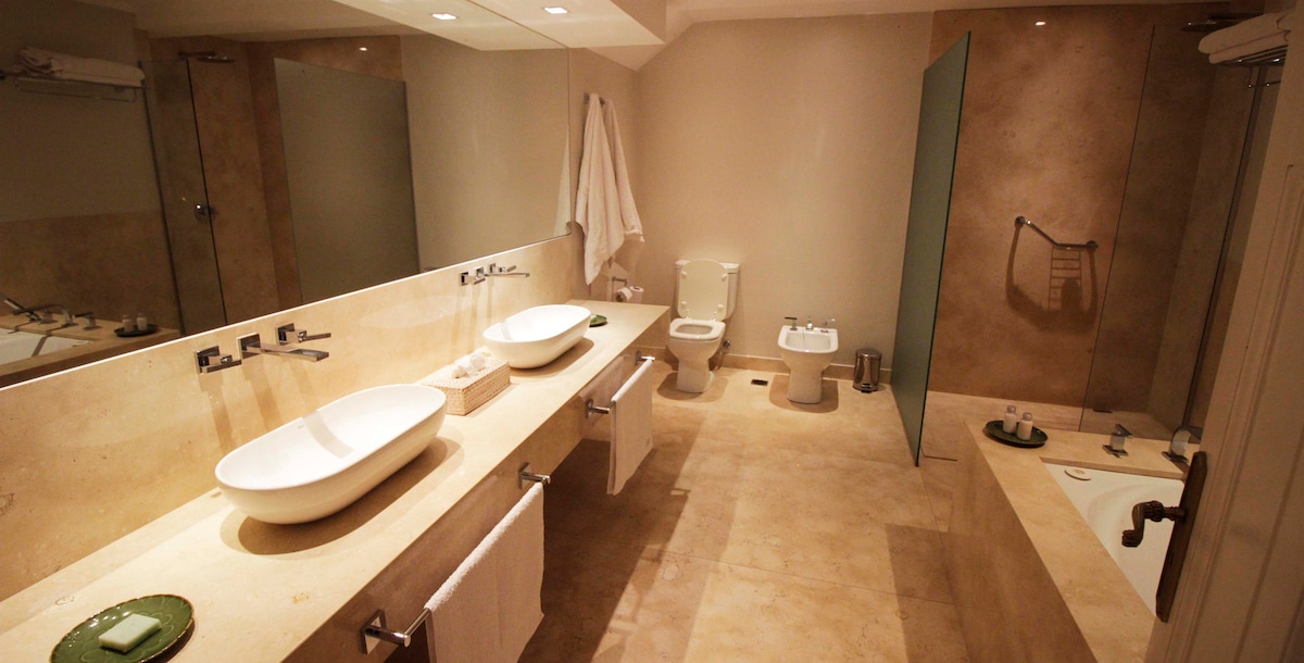 A spacious bathroom features dual vessel sinks positioned on a long, smooth countertop. Natural light enhances the neutral tones of the walls and floor. A large glass shower is situated beside a deep soaking tub, with soft towels hanging nearby and minimal decorative elements.