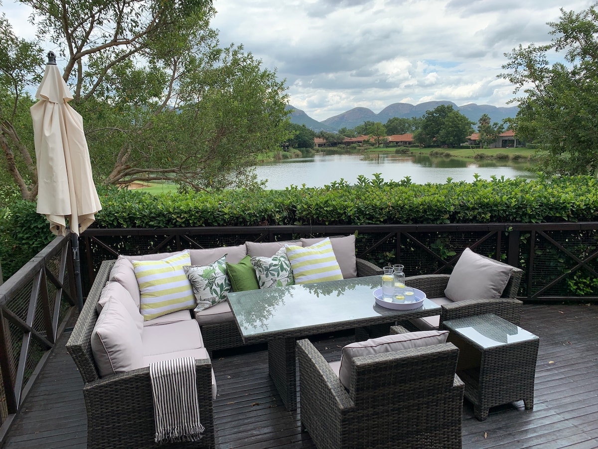 An outdoor seating area features a glass-top table surrounded by comfortable couches adorned with decorative pillows. Lush greenery frames the space, with views of a serene lake and mountains in the distance. An umbrella provides shade for the seating area.