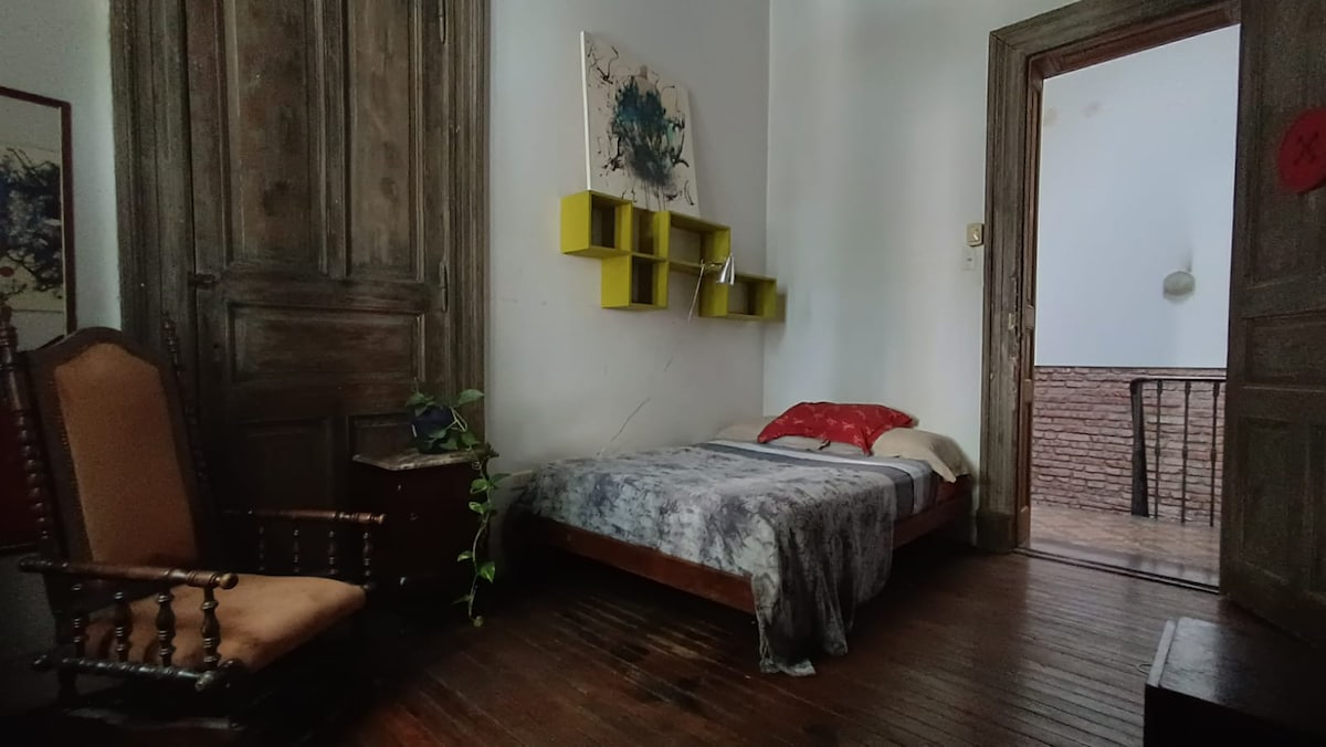 A private room is furnished with a bed draped in a patterned blanket. A wooden chair is positioned beside the bed, and a modern shelving unit is mounted on the wall featuring green accents. Natural light enters through a nearby door leading to a balcony.