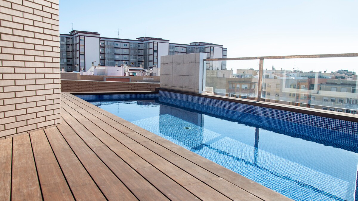 A private terrace features a tiled swimming pool framed by wooden decking. The expansive area offers unobstructed views of the surrounding cityscape, complemented by clear blue skies. Tall buildings are visible in the distance, providing a sense of the urban environment.